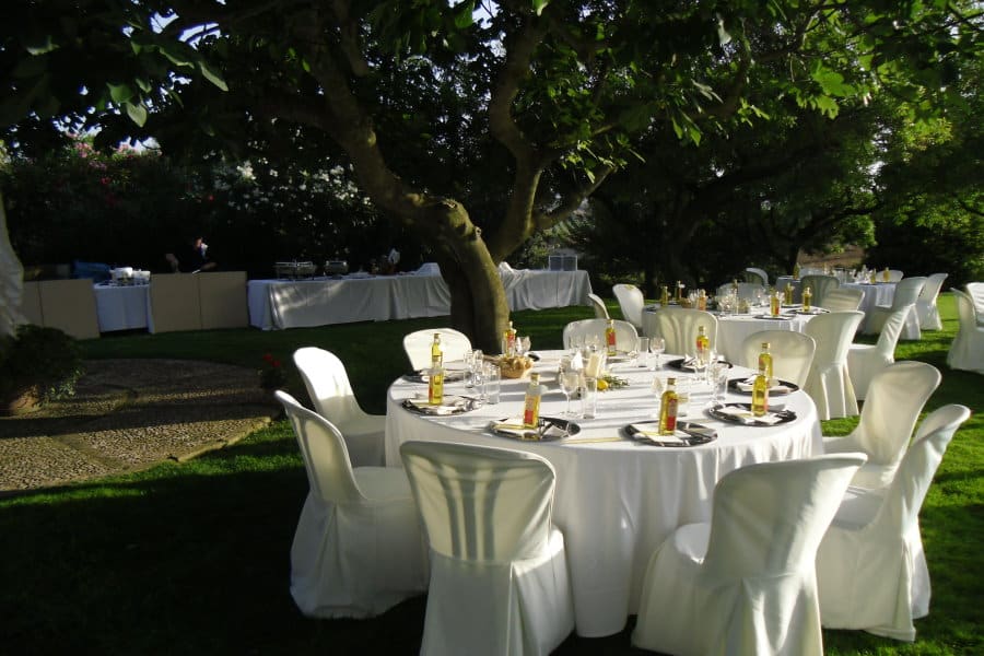 Essbereich im Freien mit Catering Mallorca unter einem Baum mit weiß bezogenen Stühlen und gedeckten Tischen für eine Mahlzeit.