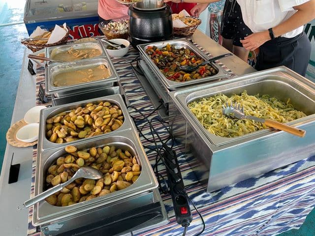 Un buffet con bandejas de comida, que incluyen patatas asadas, pasta y verduras, sobre una mesa con personas sirviéndose ellas mismas en un evento de catering en Mallorca.